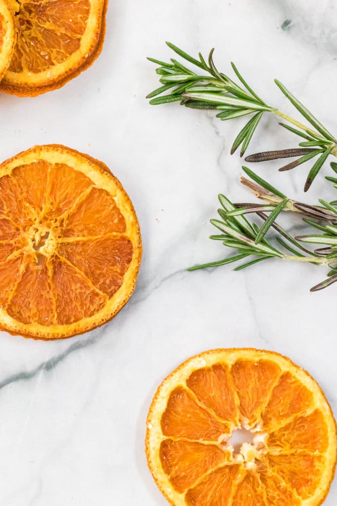 Drehydrated orange slices spread out across a white background with sprigs of fresh rosemary in the background