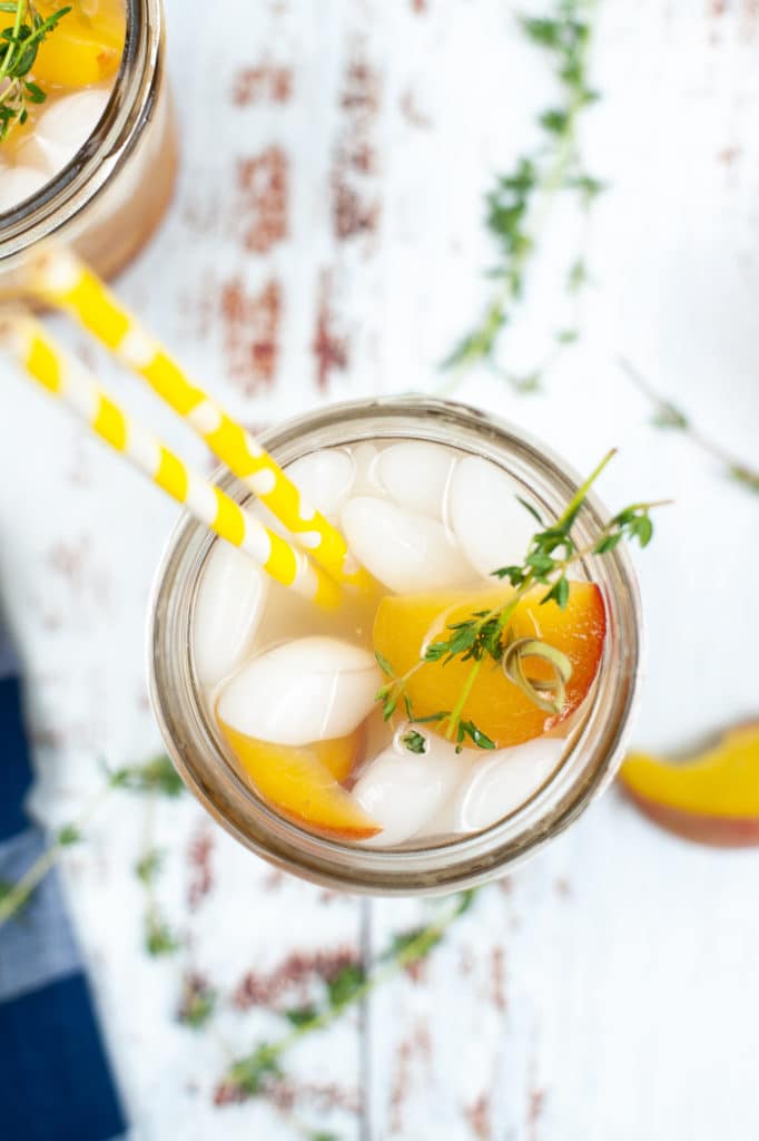 overhead shot of a gin peach cocktail served in a glass jar with slices of fresh peach, ice and a sprig of fresh thyme