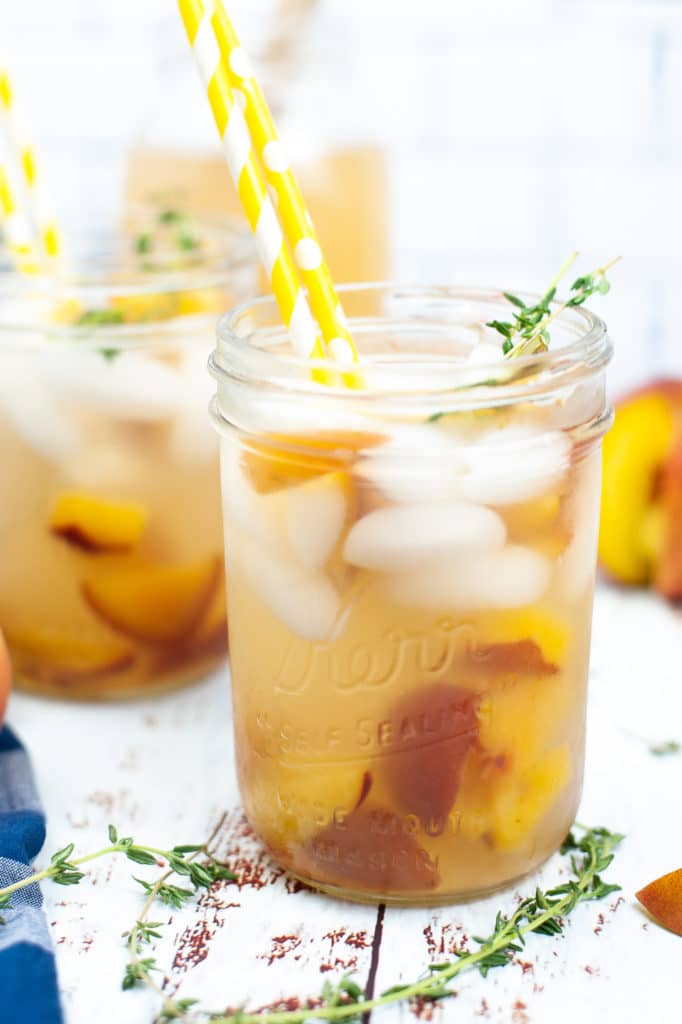 Front shot of a peach gin cocktail being served in a mason jar loaded with ice and a sprig of fresh thyme, which a yellow straw