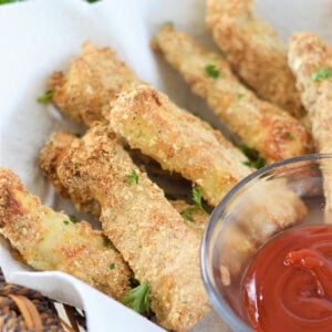 Overhead photo of baked fish fingers in a serving basket lined with parchment paper with a bowl of tomato sauce beside it