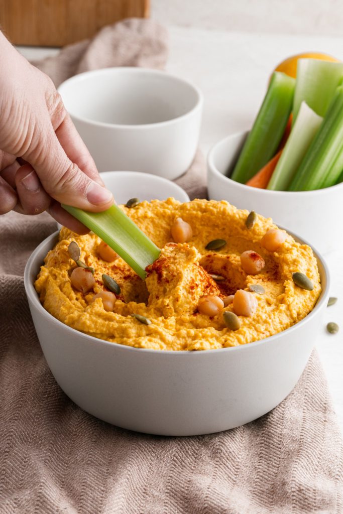 Celery sticks dipping into Roasted Garlic and Pumpkin Hummus served in a white bowl