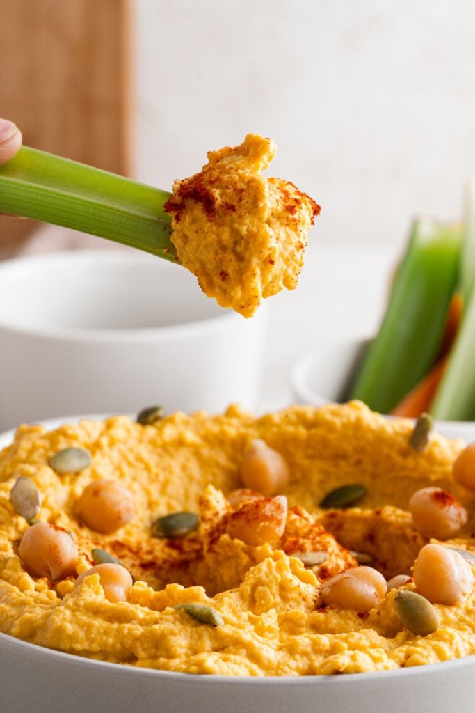 Celery sticks dipping into Roasted Garlic and Pumpkin Hummus served in a white bowl