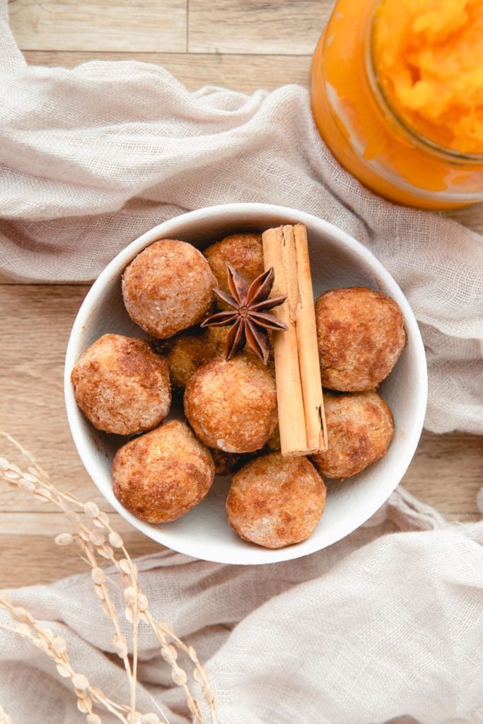 Pumpkin Protein Balls piled in a white bowl and topped with cinnamon sticks and star anise with a jar of pumpkin puree in the background