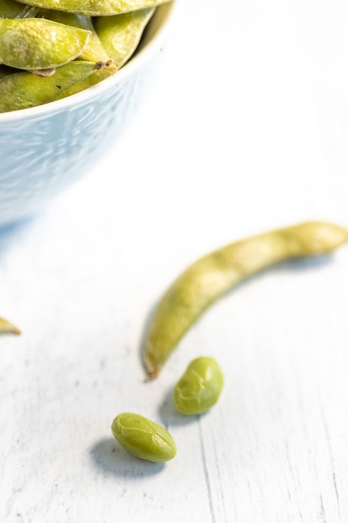 Shelled edamame beans lying beside the pod against a white background