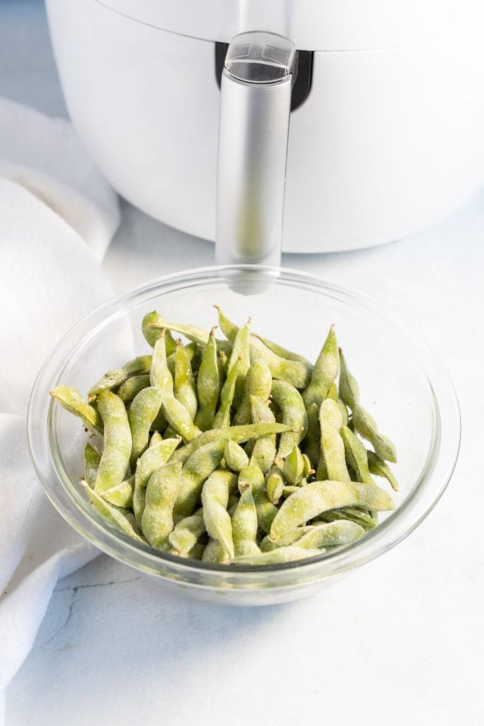 Frozen edamame in a small glass bowl in front of a white air fryer