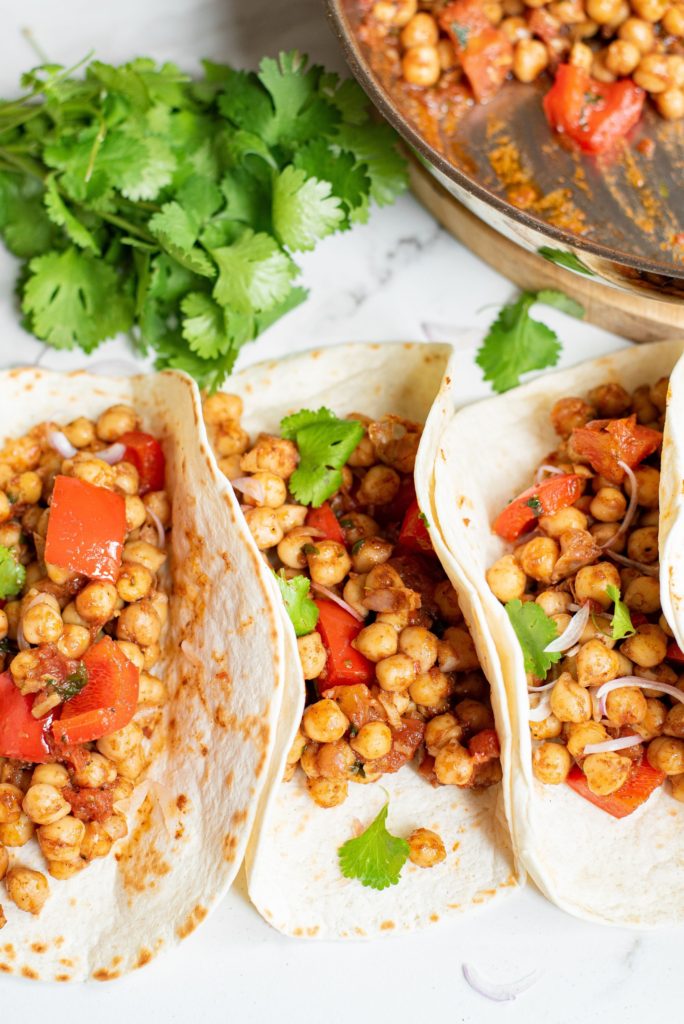 A row of chickpea tacos in flour tortillas against a background of cilantro and chickpea filling