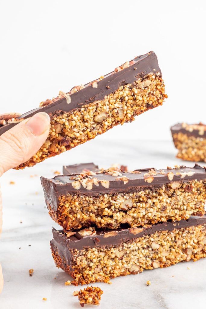 Hand lifting a chocolate quinoa bar from a stack against a white background