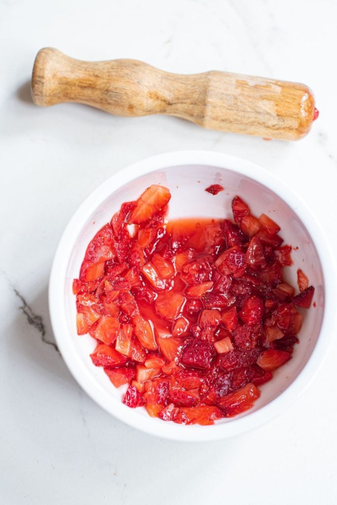 A white bowl of crushed strawberries with a wooden muddler on the side