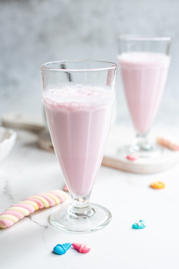 A tall glass ¾ full of a pink milkshake with a bowl of whipped cream in the background about to be added