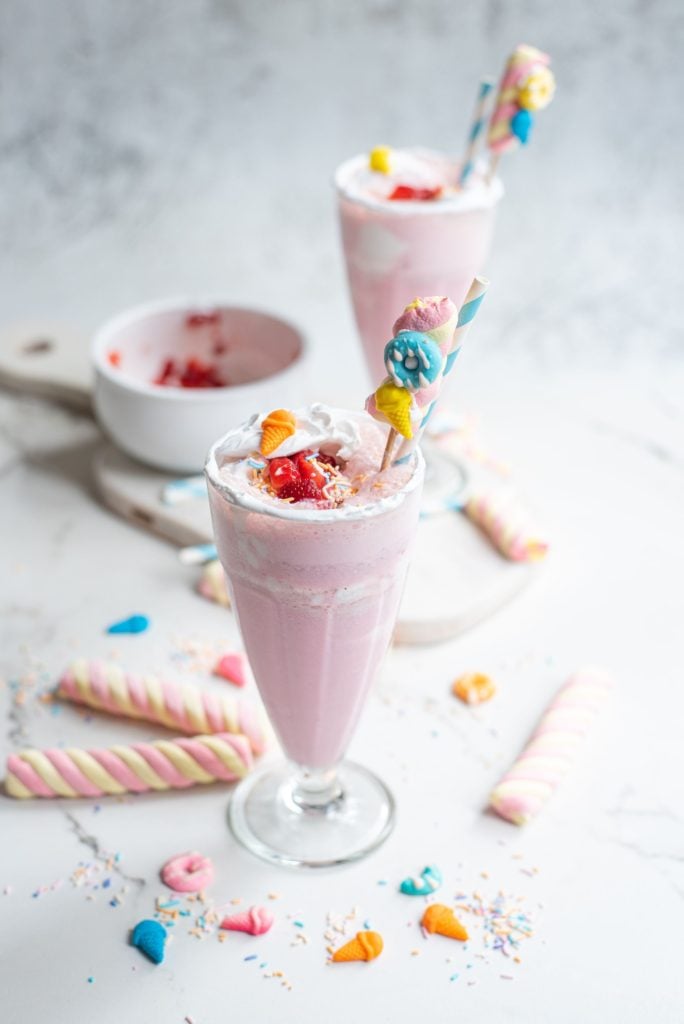 A tall glass filled with a homemade milkshake with lots of lollies for decorating sprinkled in the background
