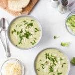 Overhead shot of a light green soup served into 2 white bowls with some cheese and bread