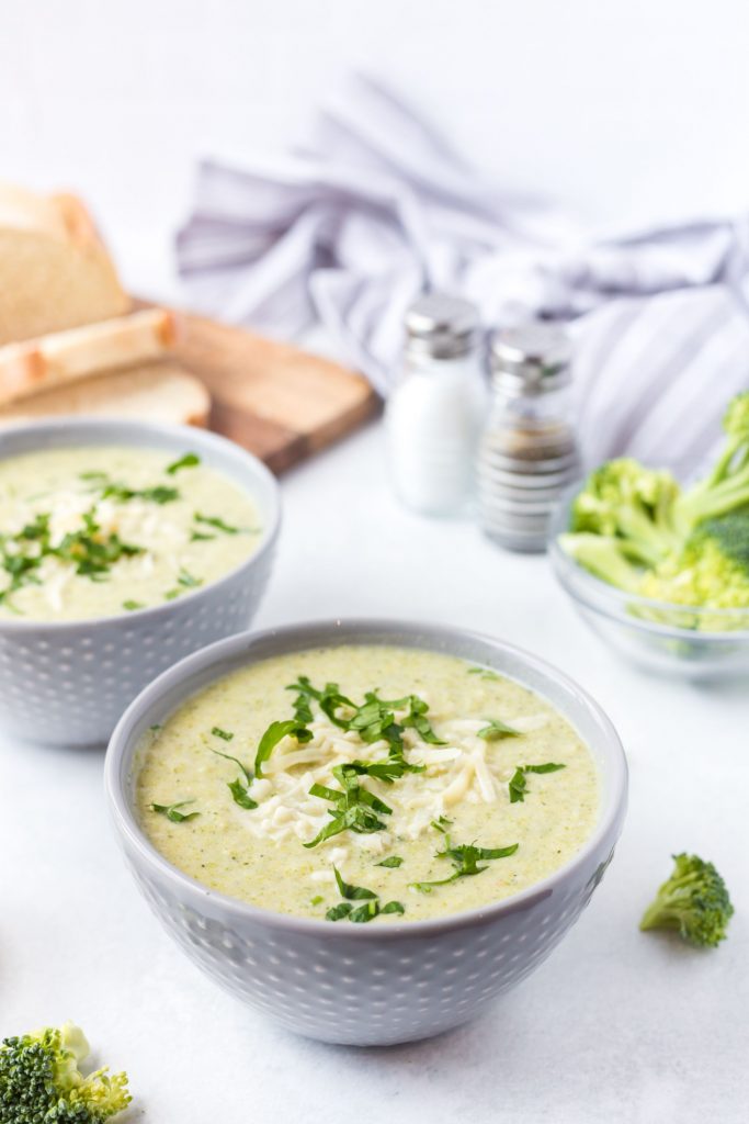Two bowls of green soup served up in white bowls with some slices of fresh bread on a chopping board in the background, and broccoli florets spread around