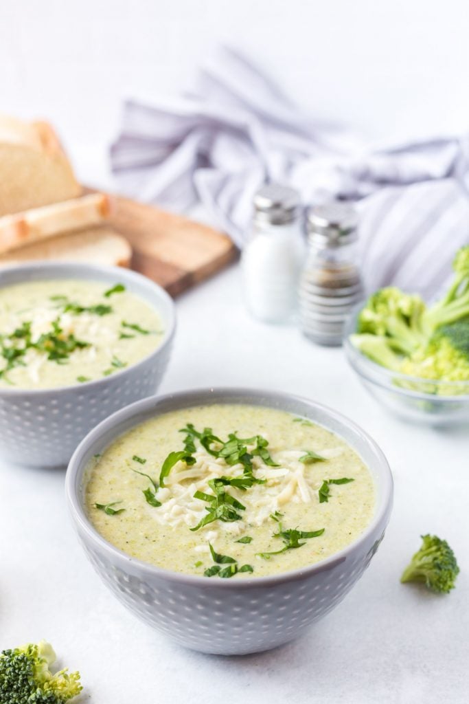 Two bowls of green soup served up in white bowls with some slices of fresh bread on a chopping board in the background, and broccoli florets spread around