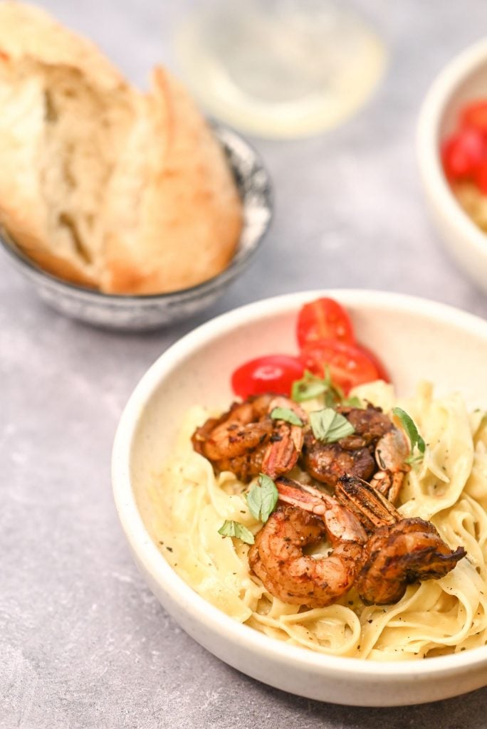 Shot of cajun shrimp alfredo pasta served in a white bowl and topped with some chopped tomatoes and basil with a loaf of crunchy bread in the background