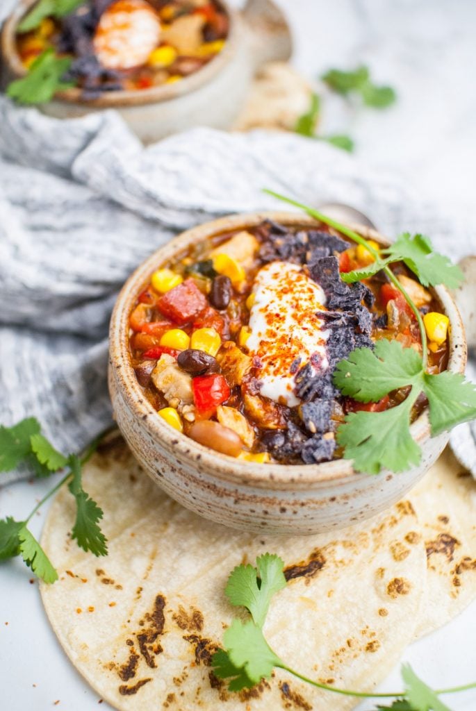 Chunky soup in a speckled brown bowl wrapped in grey linen. The soup is topped with a big dollop of sour cream and fresh cilantro