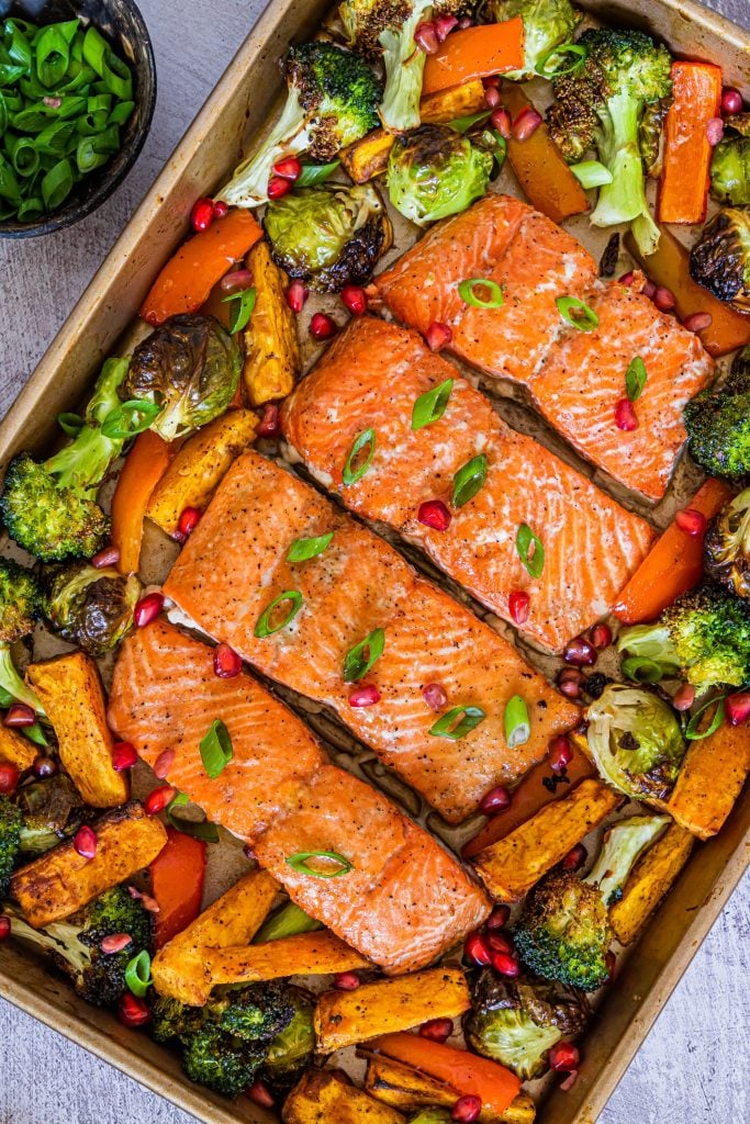 4 pieces of cooked salmon with broccoli, brussels sprouts and sweet potato in the background and sprinkled with spring onions and pomegranate