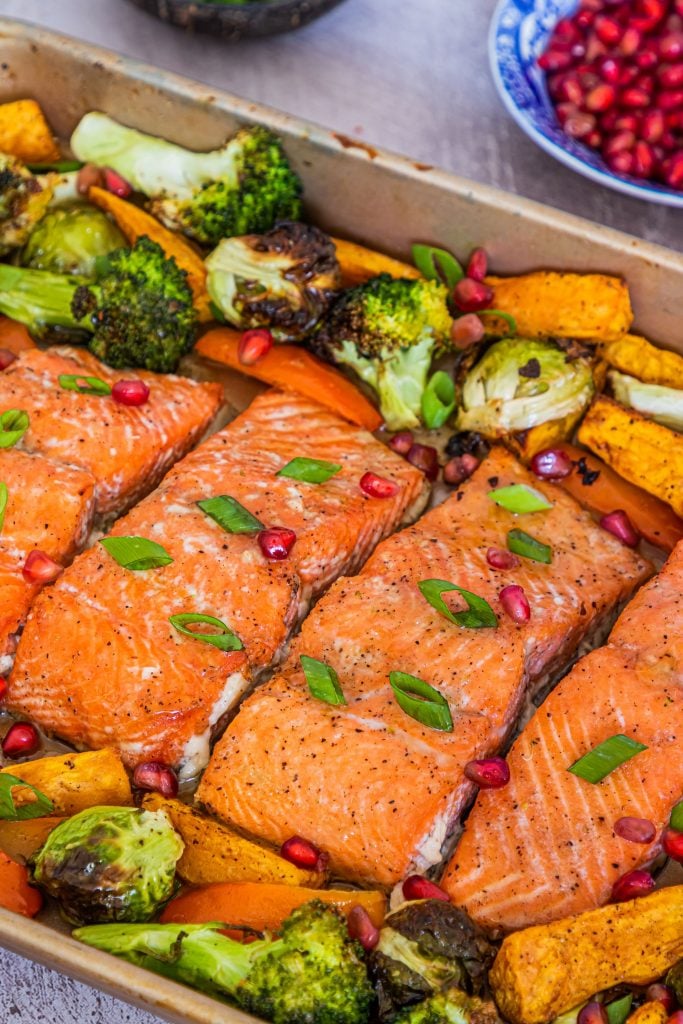 4 pieces of cooked salmon with broccoli, brussels sprouts and sweet potato in the background and sprinkled with spring onions and pomegranate with a bowl of extra pomegranate