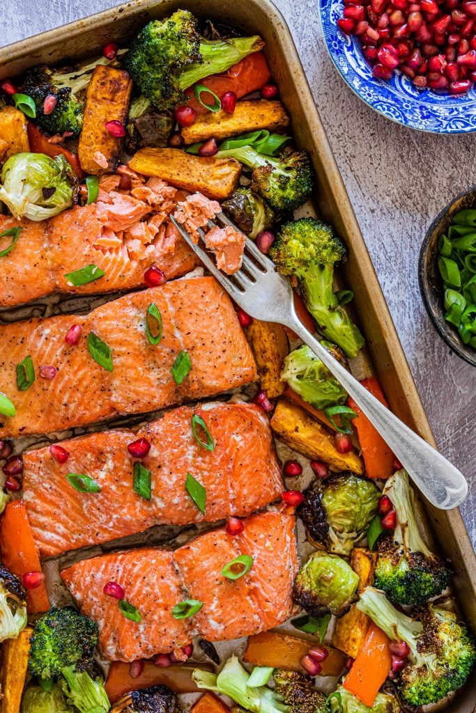 Salmon broccoli and sweet potatoes on a baking sheet with a fork pulling the salmon apart