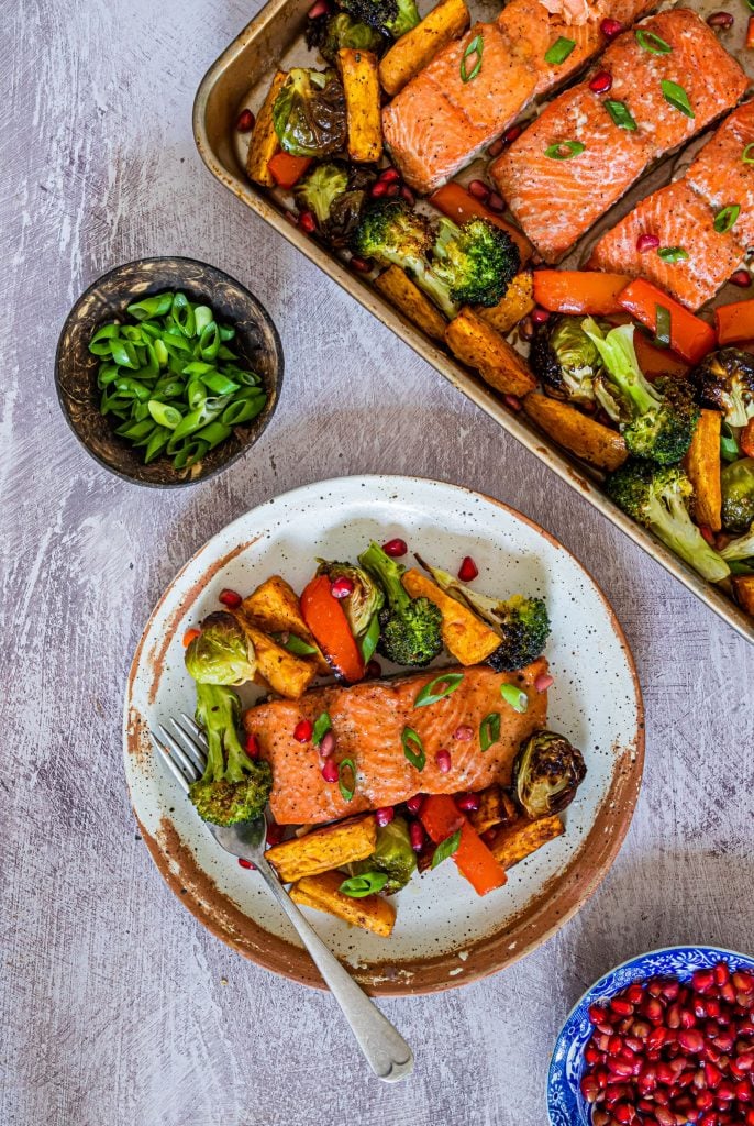 Salmon and veggies cooked on a sheet pan and served up on an off-white plate with some spring onions and pomegranate