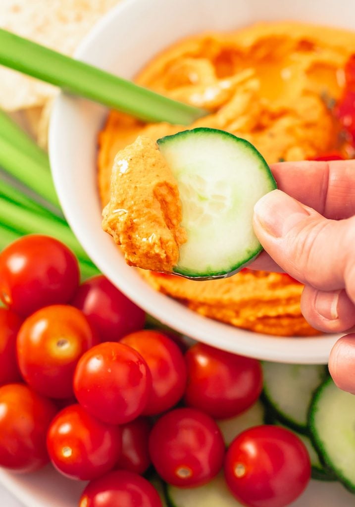 A slice of cucumber scooping up some hummus from a white bowl surrounding by other chopped veggies and baby tomatoes