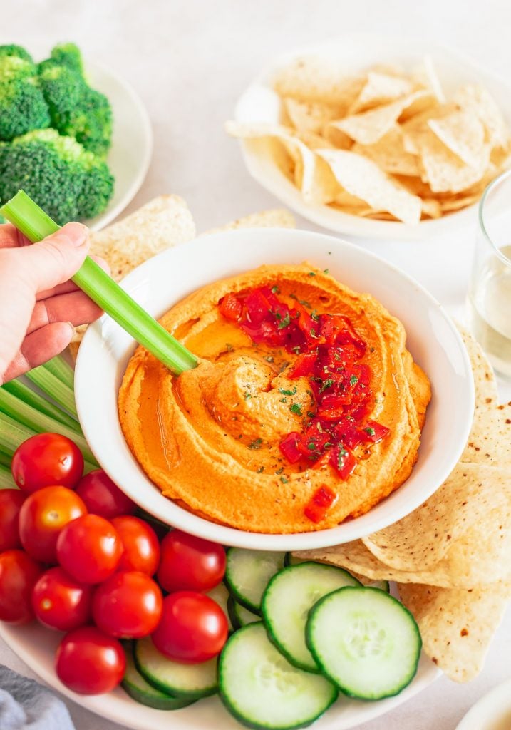 A bowl of orange hummus served in a white bowl surrounded by chopped veggies