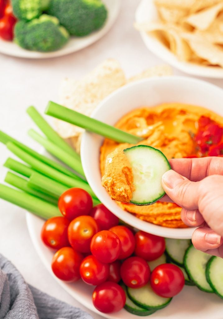 Hand using some chopped up veggies to scoop a red dip from the white bowl.