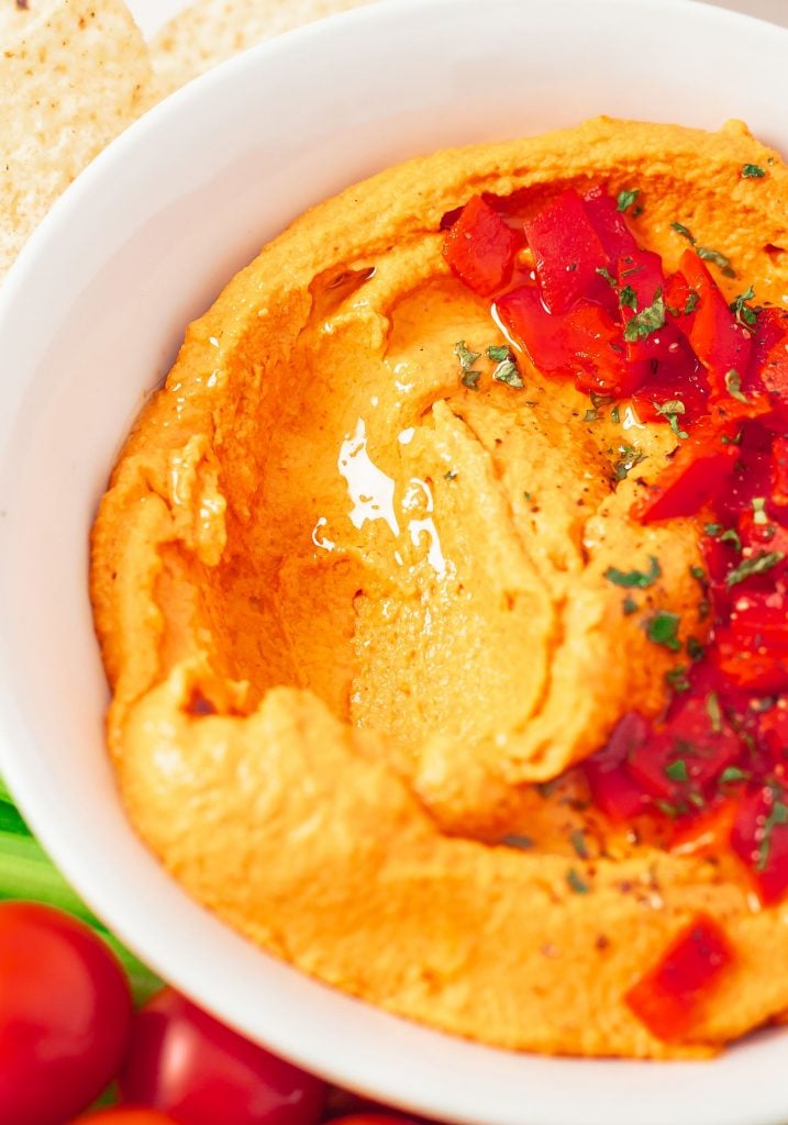 up close shot of red pepper hummus served in a white bowl with chopped red peppers and olive oil on top