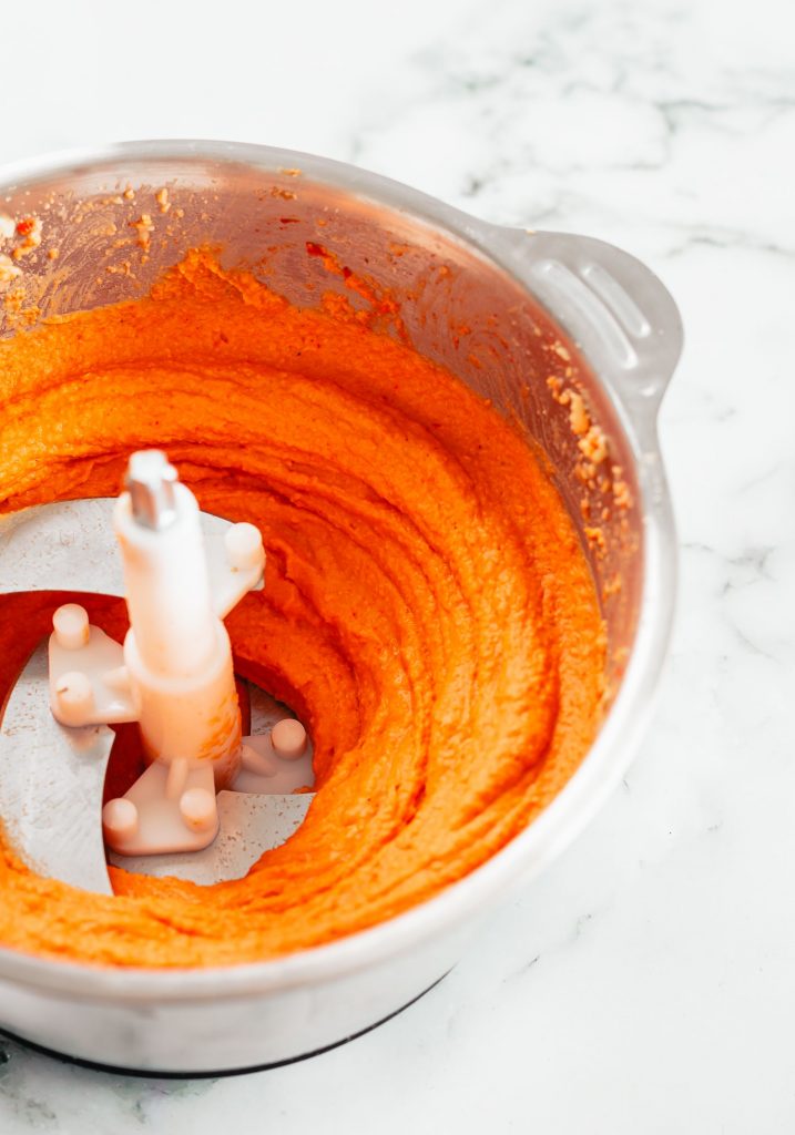 A bright orange/red hummus in the bowl of a food processor