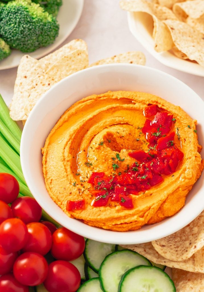 Red pepper hummus served in a small white bowl and surrounded by lots of chopped vegetables