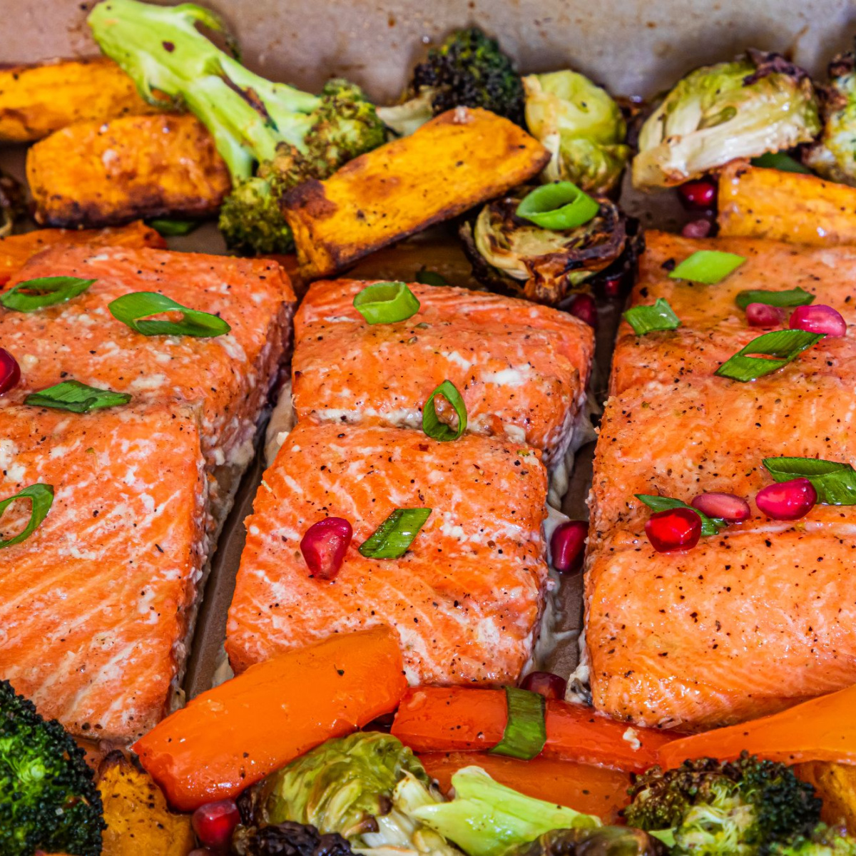 cooked salmon with broccoli, brussels sprouts and sweet potato in the background and sprinkled with spring onions and pomegranate