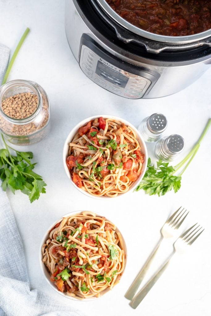 Two bowls of veggie bolognese and spaghetti in front of an instant pot containing extra sauce with lots of fresh parsley in the background