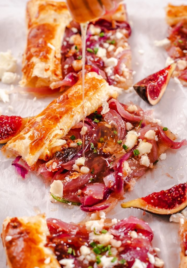 Puff pastry caraemlized onion tart being drizzled with maple syrup on some parchment paper with wedges of fresh figs surrounding