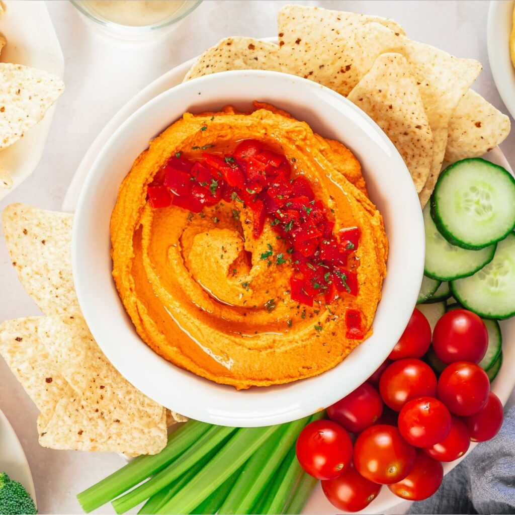 A bowl of orange hummus served in a white bowl surrounded by chopped veggies