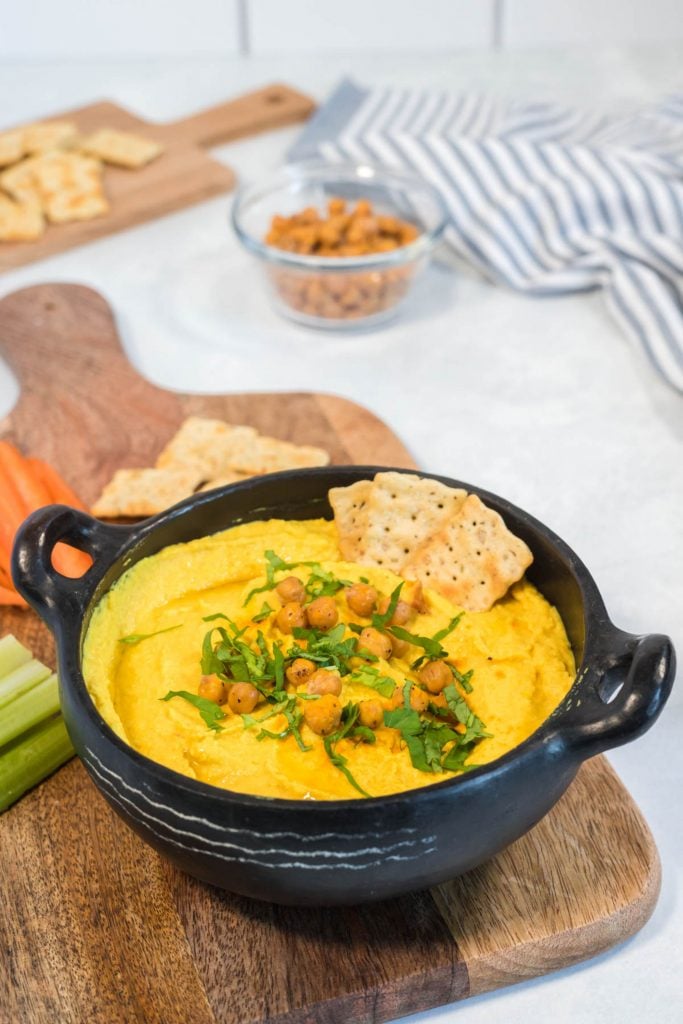 Golden hummus served in a black bowl and topped with chickpeas and chopped parsley. There are veggie sticks and crackers in the background