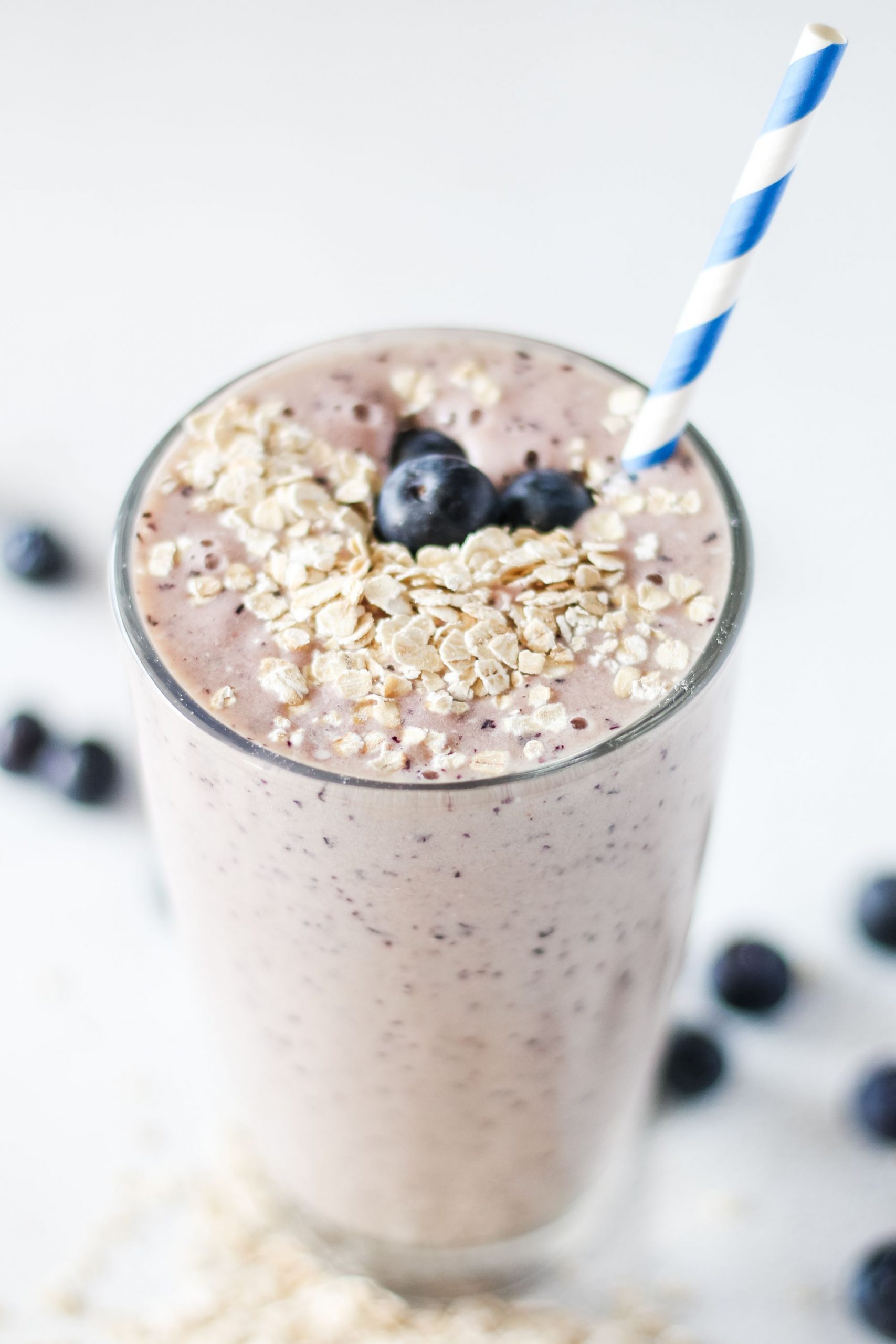 Light pink smoothie in a tall glass with a straw served with some fresh blueberries and oats on top
