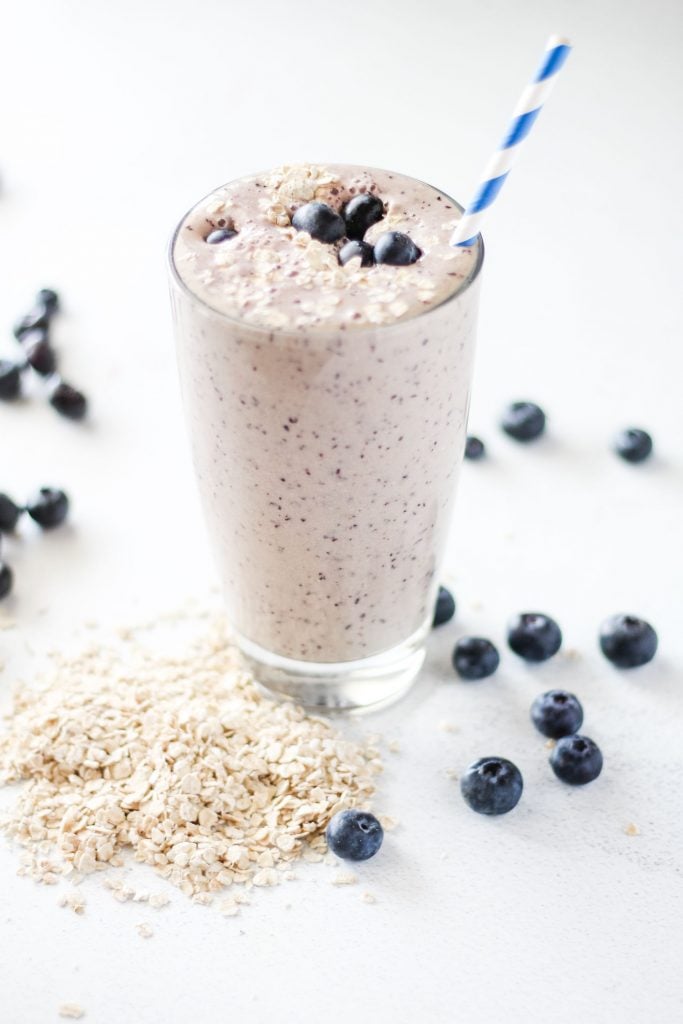 front shot of a banana and blueberry smoothie served in a tall glass with a straw. Topped with blueberries and oats which are also in the background