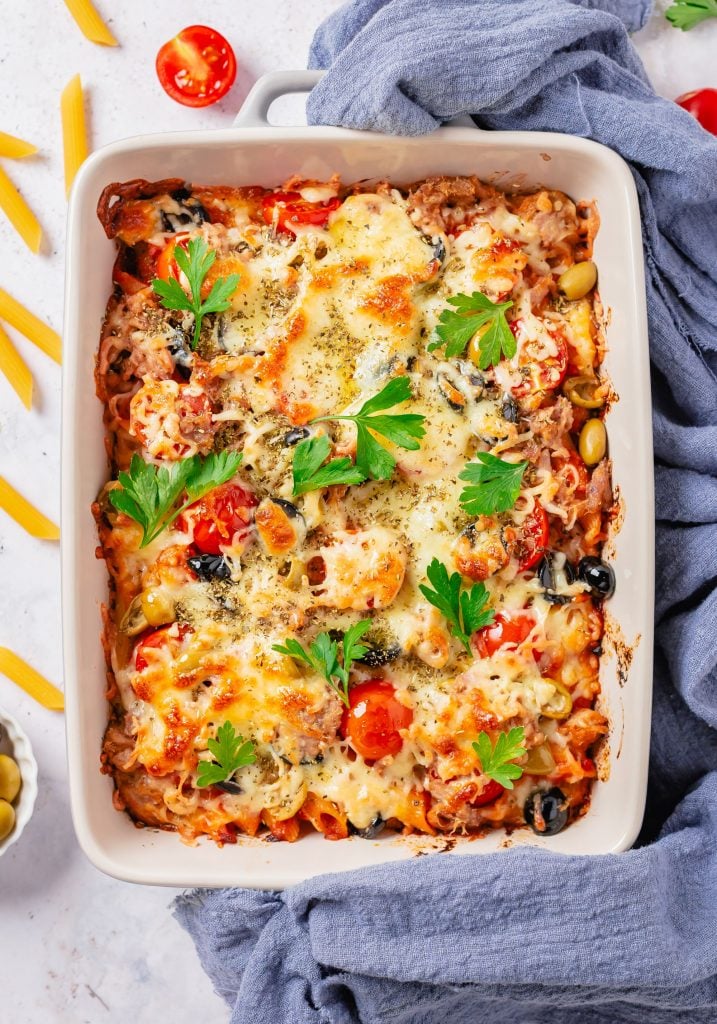 Overhead shot of healthy tuna casserole served in a white casserole dish and surrounded by dry pasta and cherry tomatoes