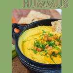 overhead shot of turmeric hummus served in a black bowl with a side of veggie sticks and crackers
