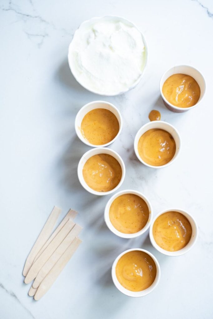 Mango puree poured to the halfway mark of a popsicle mold or small espresso cup. the coconut cream is in a bowl beside the cups, as are the wooden icy pole sticks