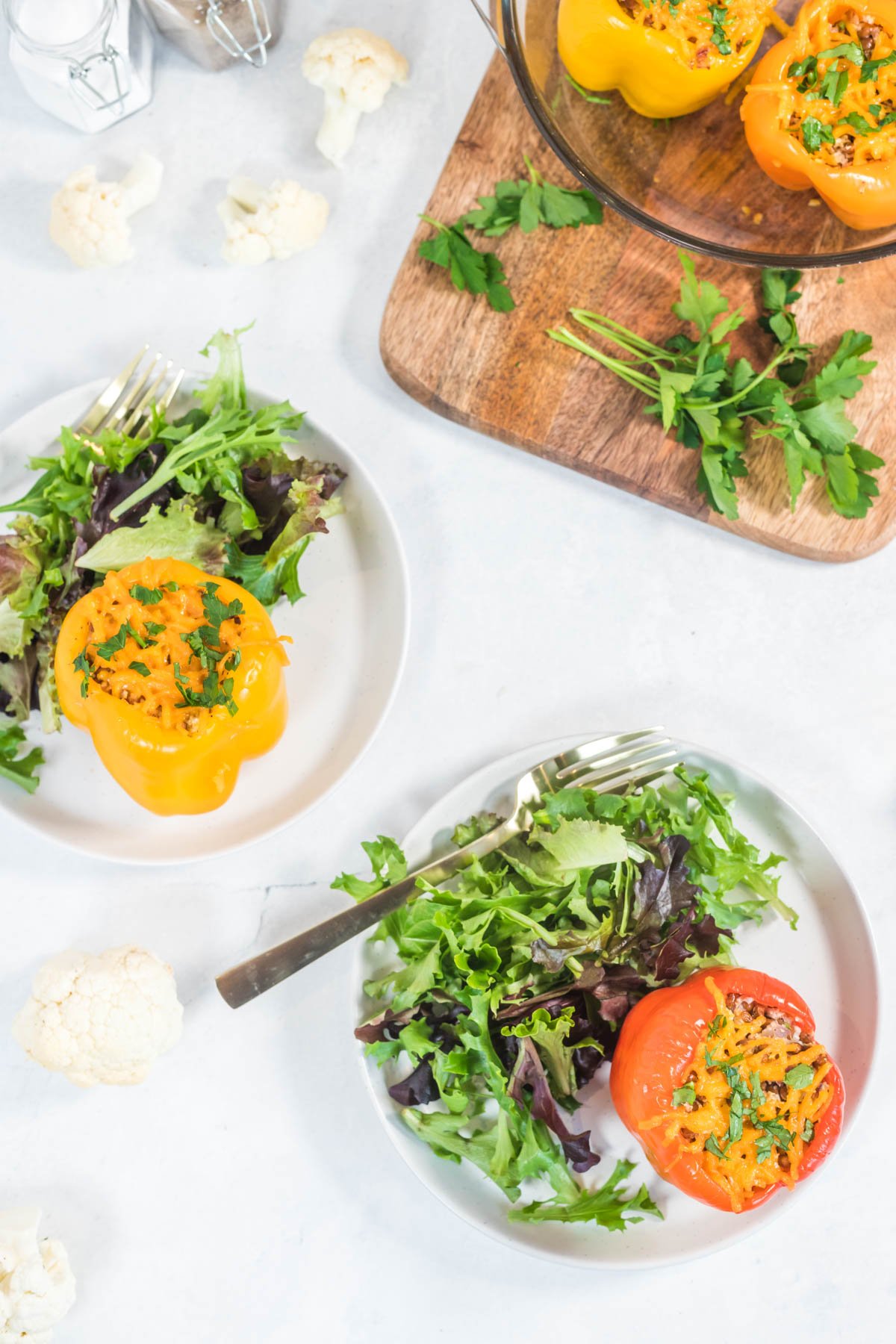 2 different colored bell peppers stuffed with cauliflower rice and herbs, served on white plates with a simple side salad