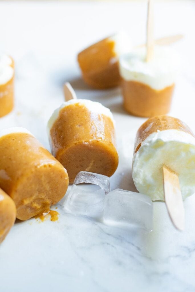 6 popsicles with an orange layer and a white layer sitting on a bench amongst ice cubes