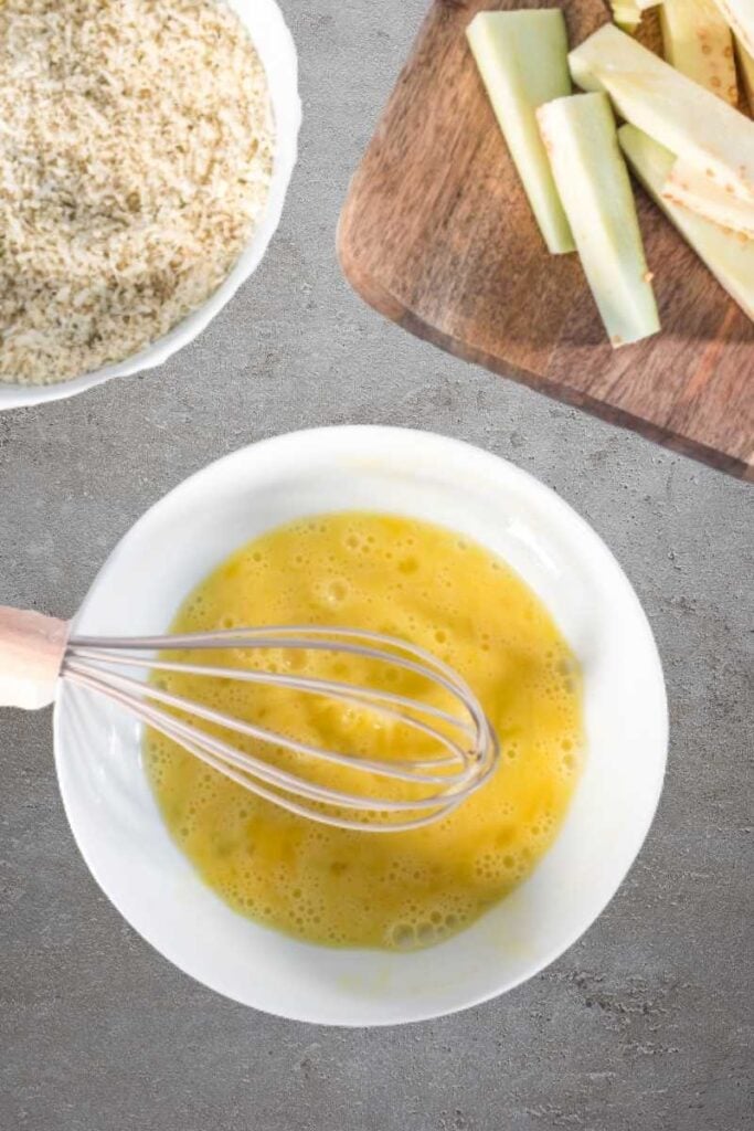 An egg being whisked in a white bowl. There are some uncooked eggplant fries and panko breadcrumbs in the background