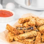 A stack of crispy eggplant fries served on some parchment paper with tomato sauce and an air fryer in the background