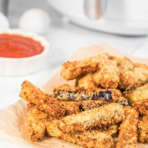 A stack of crispy eggplant fries served on some parchment paper with tomato sauce and an air fryer in the background