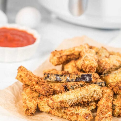 A stack of crispy eggplant fries served on some parchment paper with tomato sauce and an air fryer in the background