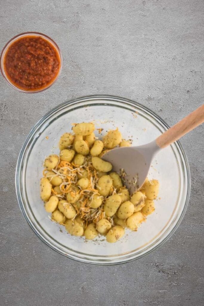 A large glass mixing bowl containing gnocchi, cheese and seasonings