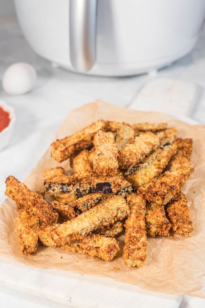 A stack of panko crumbed eggplant fries served on parchment paper with an air fryer in the background