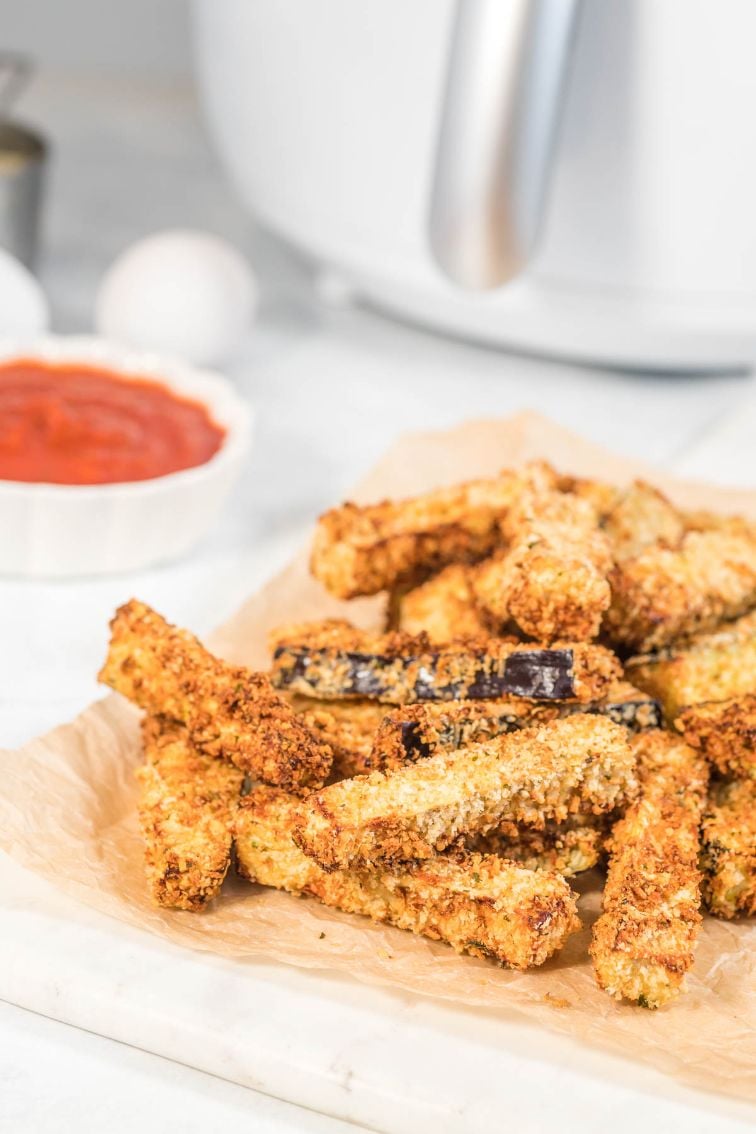 A stack of crispy eggplant fries served on some parchment paper with tomato sauce and an air fryer in the background