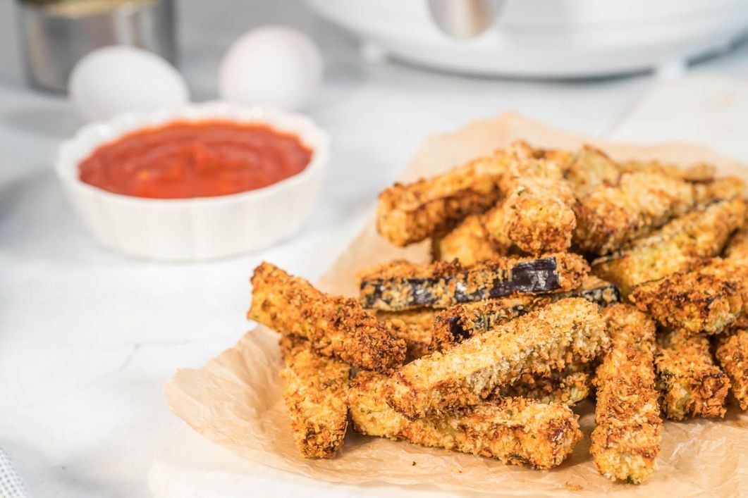 air fryer eggplant fries stacked on some parchment paper with a side of tomato sauce