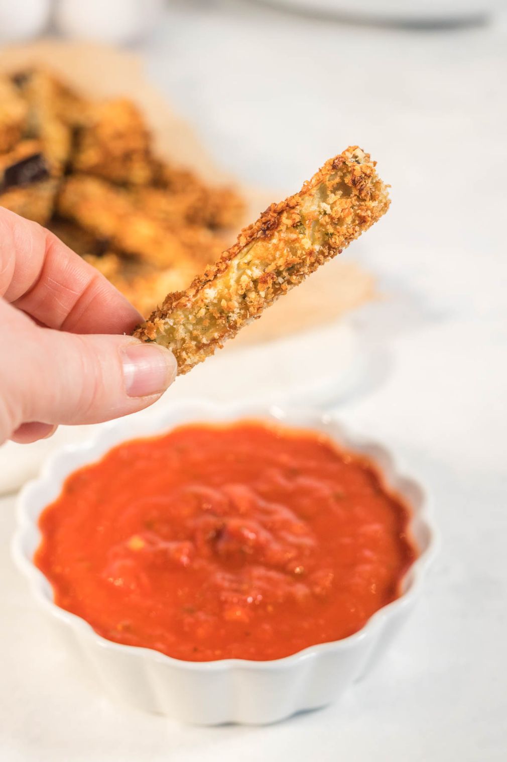 An eggplant fry being held over a small bowl of tomato sauce